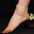 A foot adorned with the Unisex Bracelet, crafted with 14k gold-filled accents and featuring colorful circular charms, including a Turkish Evil Eye for spiritual protection. White flowers bloom gracefully in the background.